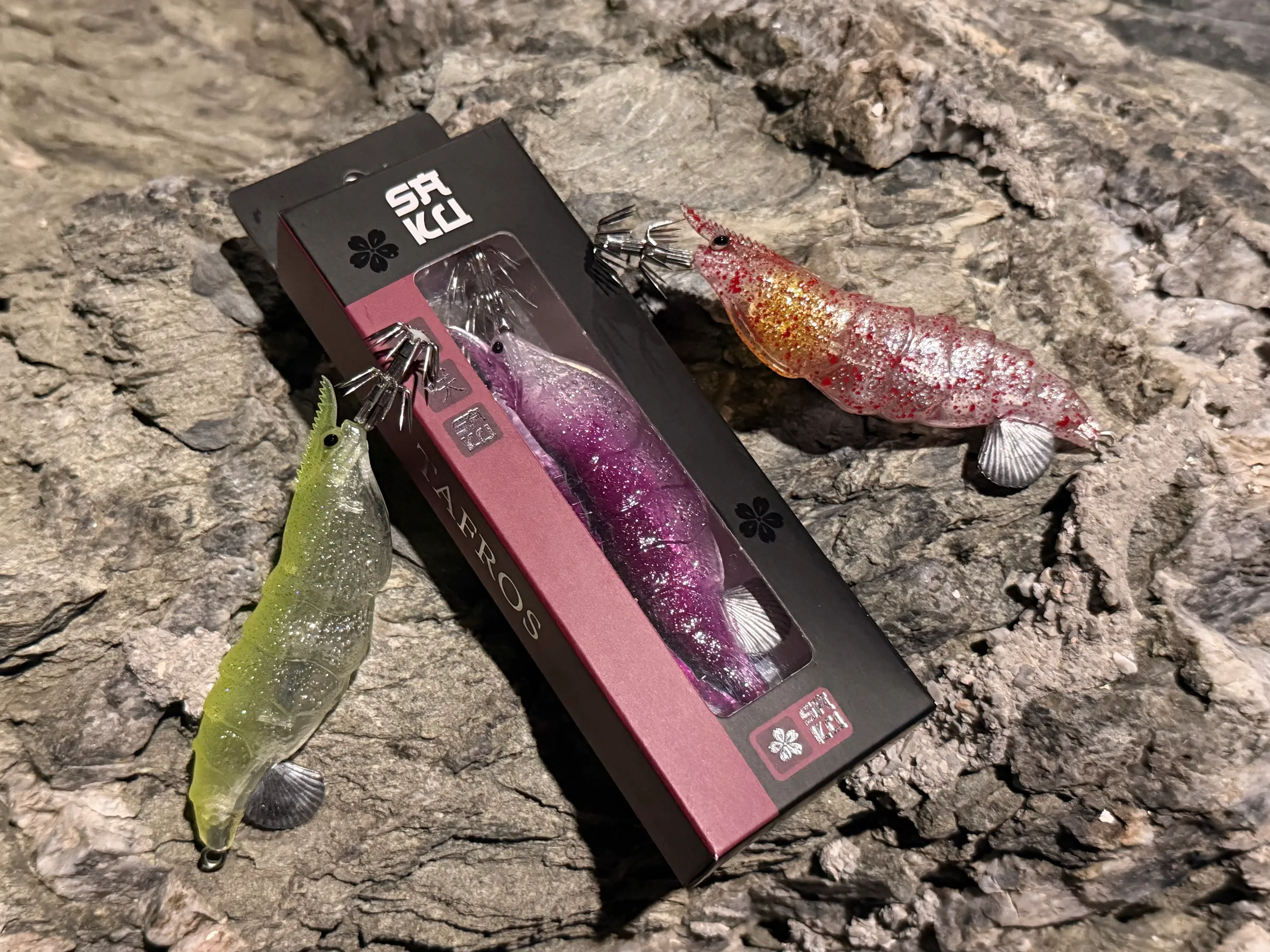 TAFROS squid jigs in chartreuse and red speckle beside the violet jig in its retail box on rock