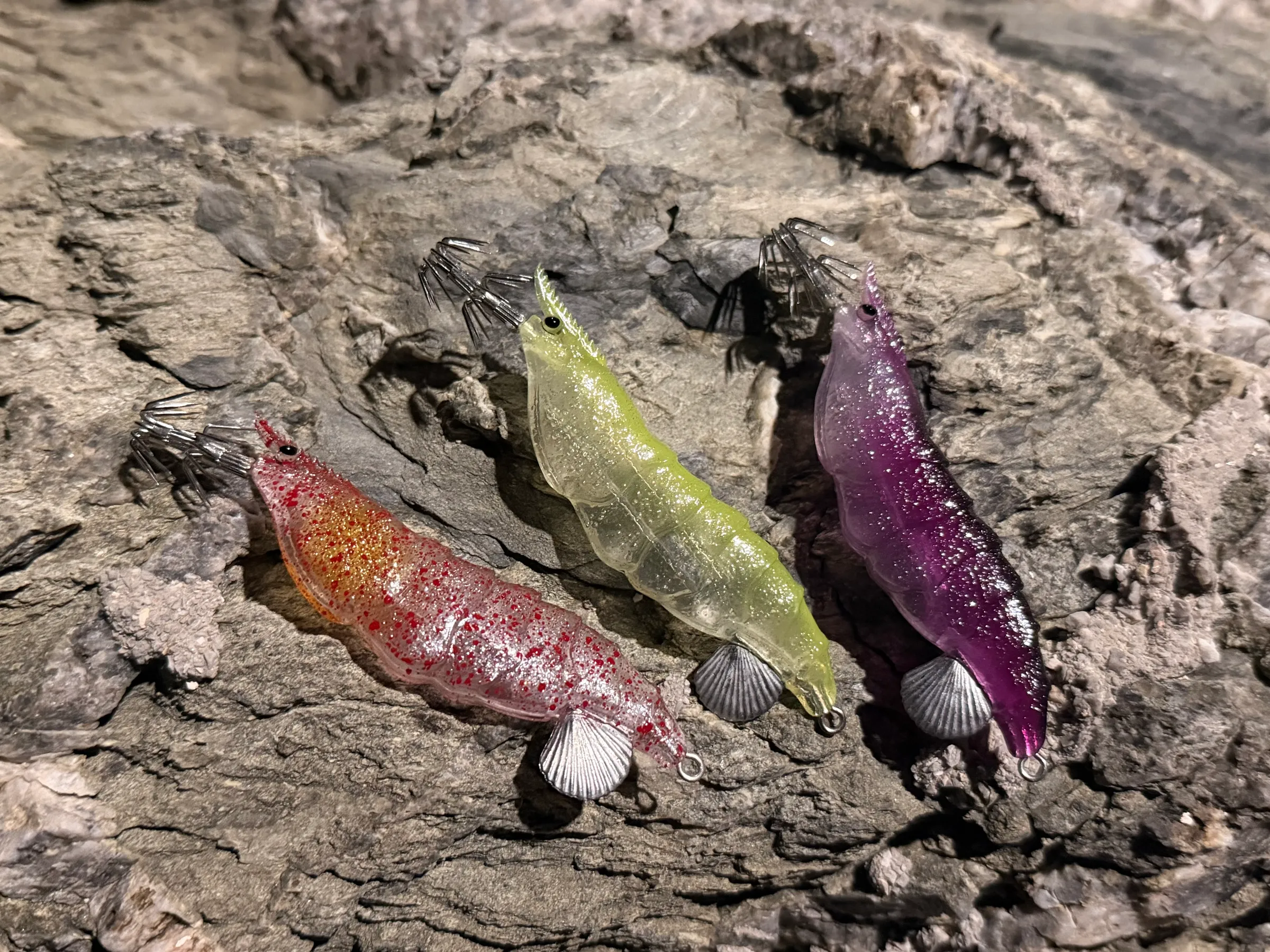 All three TAFROS squid jig colours resting on dark volcanic rock under low side light